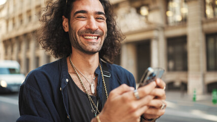 Young attractive italian guy with long curly hair and stubble is using mobile phone. Stylish man...