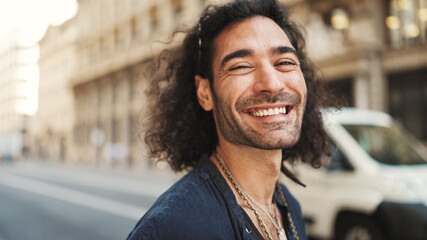 Young italian guy with long curly hair and stubble stands on the sidewalk next to the road, looks around and smiles