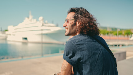 Young italian guy with long curly hair and stubble sits in harbor and looks at ships and yachts