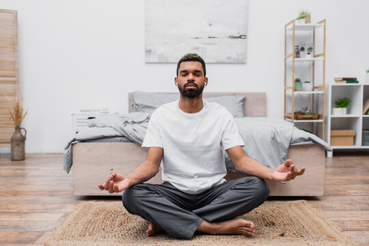 bearded african american man in wireless earphone meditating with closed eyes while sitting on rug near bed.