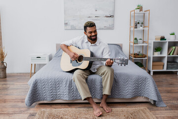 barefoot and happy african american man with dyed hair and beard playing acoustic guitar in bedroom.