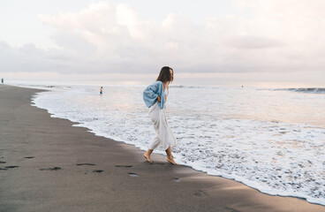 Gorgeous Caucasian woman enjoying vacations while walking on the beach feeling calm and carefree. Beautiful healthy female in white summer cloth strolls on seashore during holidays travel