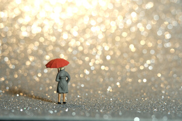 Miniature people toy figure photography. A lady wearing raincoat and umbrella, standing enjoying golden yellow bokeh light background