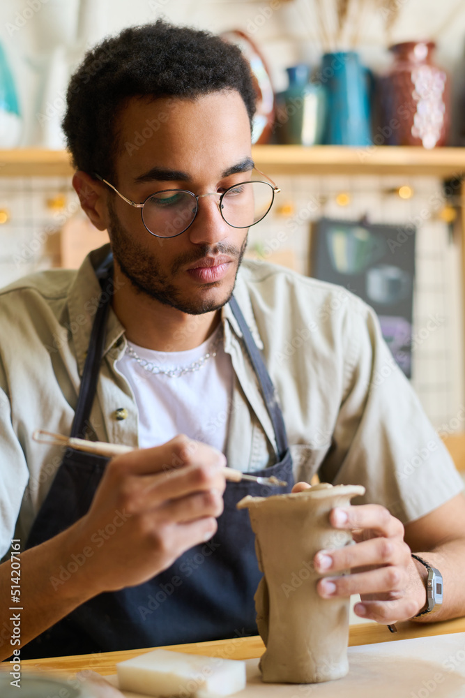 Aufkleber Serious guy in eyeglasses and apron using handtool while creating clay jug or vase for collection of earthenware items for sale