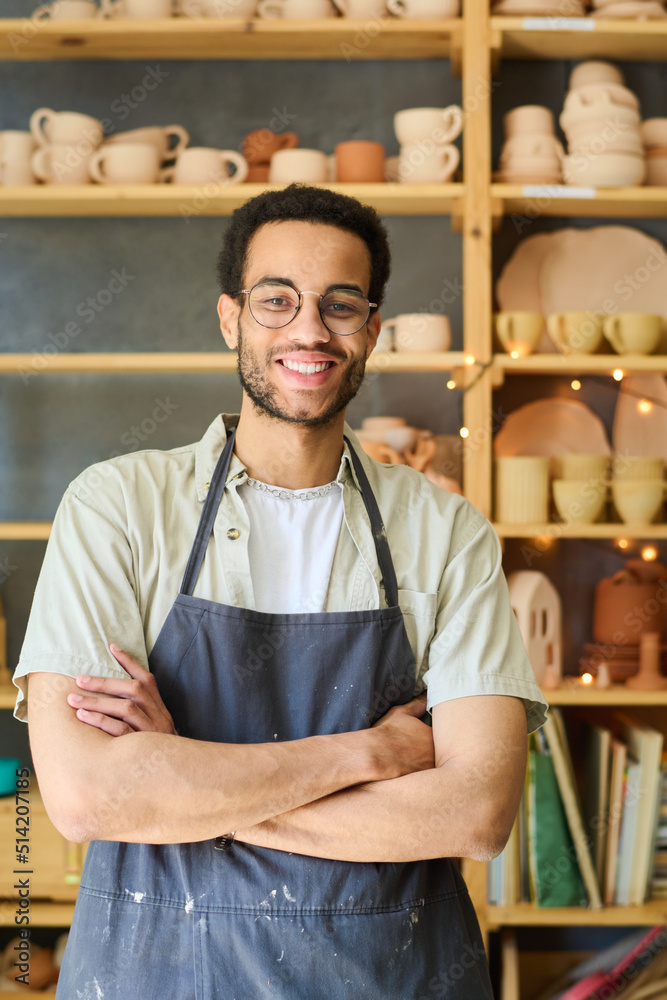 Poster young successful craftsman in apron and eyeglasses crossing arms by chest while looking at camera ag