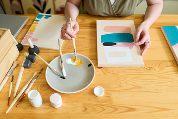 ABove angle of hands of young female artist mixing colors on plate while sitting by wooden table and painting abstract picture on paper