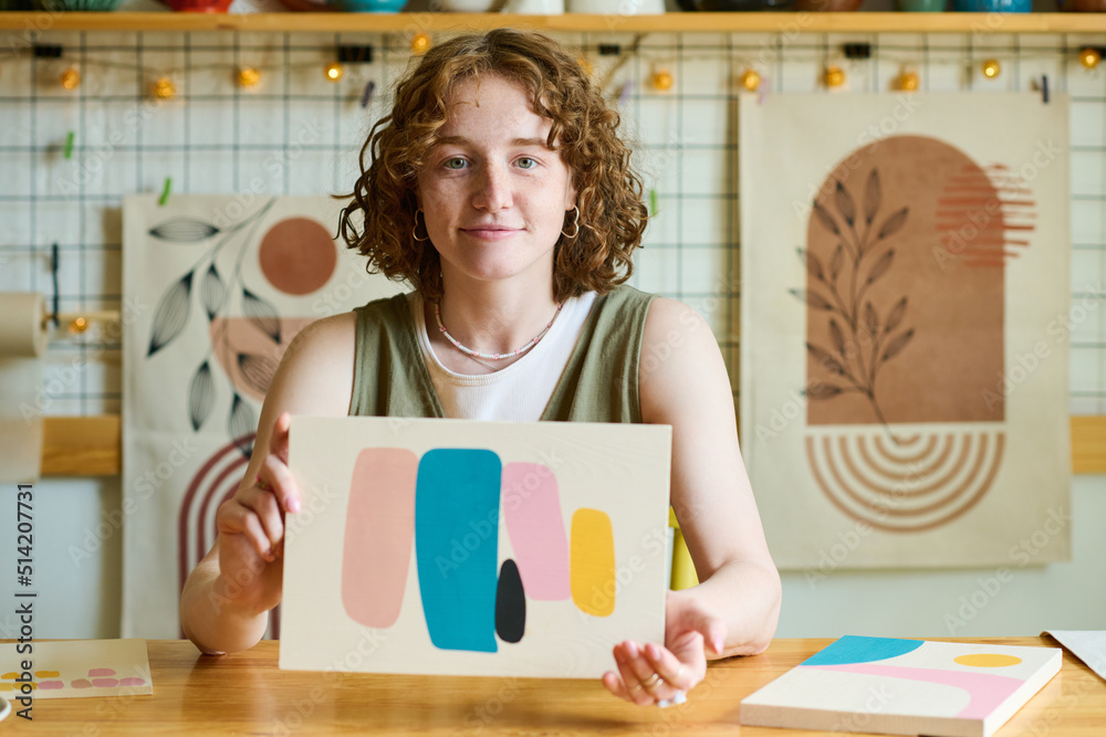 Aufkleber Young smiling female holding abstract painting created by herself while showing it to you and looking at camera in workshop or studio
