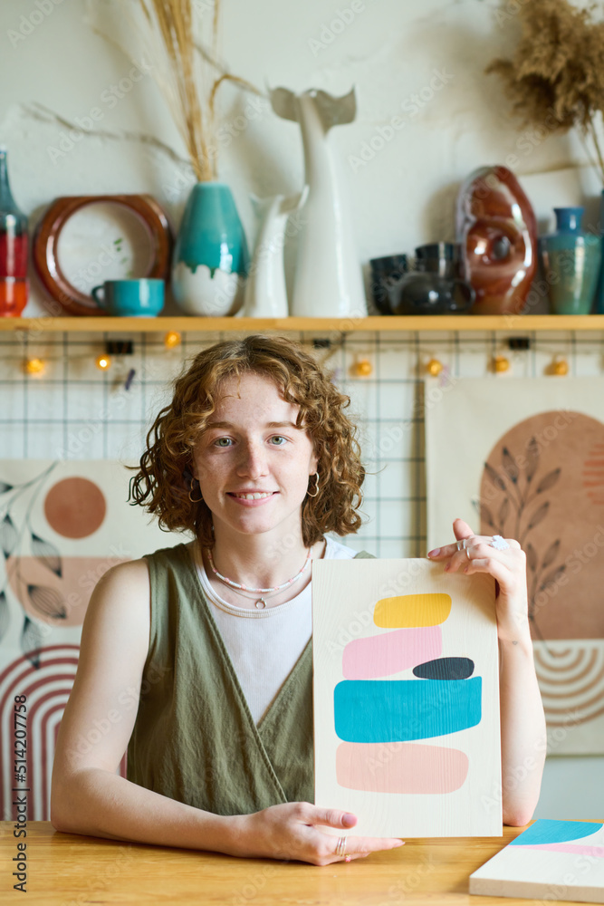 Poster young smiling woman in casualwear sitting by table and showing new abstract painting created by hers