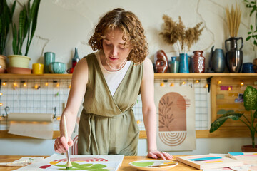 Young creative female bending over table while painting abstract picture on paper with paintbrush...