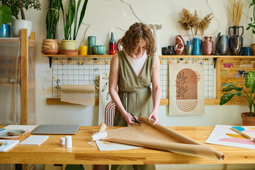 Young female clerk of souvenir shop cutting piece of wrap paper for packing gift chosen by client...