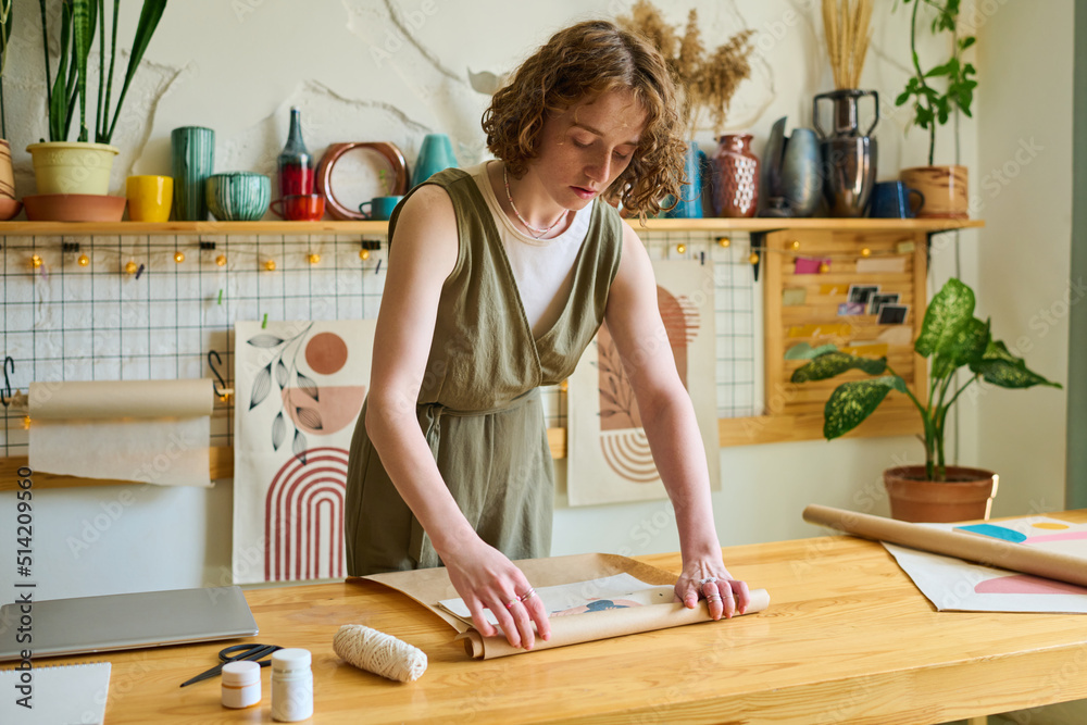Poster young female clerk bending over table while wrapping self made abstract painting into paper while pa
