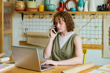 Young confident female owner of craft shop talking on mobile phone and looking through online orders of clients in front of laptop