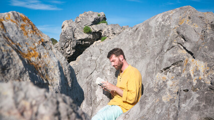 Hombre blanco barbudo leyendo un libro entre rocas en playa rocosa