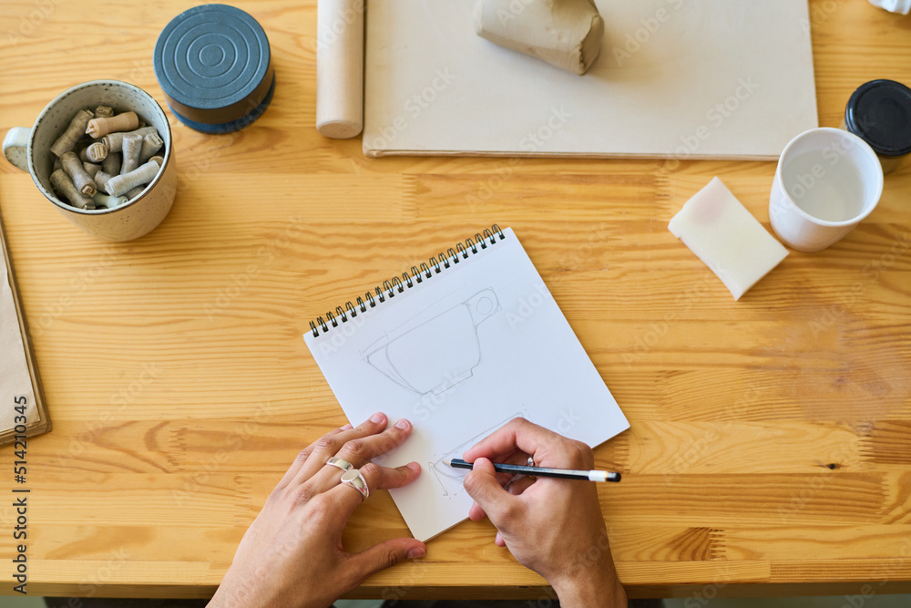 Poster above angle of hands of young craftsman or artisan drawing sketch of new earthenware item in notepad