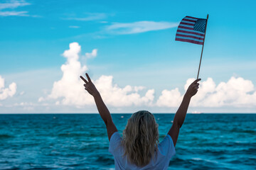 A blonde girl is standing on the ocean. In his right hand he holds an American flag, and his left hand shows 2 fingers up. Soft focus