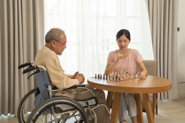 Asian daughter and old father with wheelchair, family playing a chess game together at home. People lifestyle. Activity. Therapy recover mind.