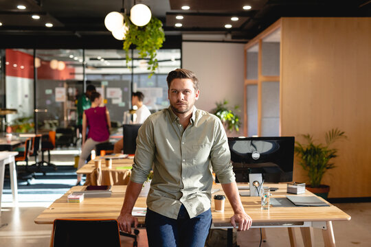 Portrait of confident caucasian young businessman leaning on desk in creative office