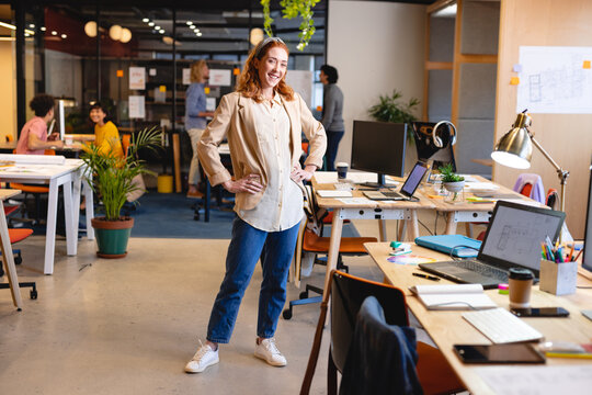 Portrait of smiling caucasian young businesswoman standing with hands on hips at workplace