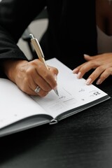 A girl sits at a black wooden desk and writes notes in her day planner and ccomputer, top view. A diary and planner notebook for planning agendas, reminders, schedules, daily appointments,  