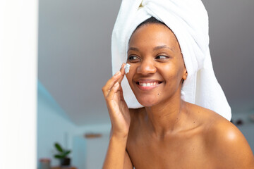 Smiling young african american woman with head wrapped in towel applying face cream, copy space