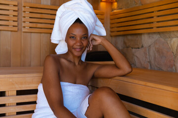Portrait of confident young african american woman wrapped in towels sitting in sauna