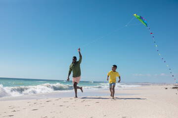 Playful african american father and son running while flying kite at beach on sunny day, copy space