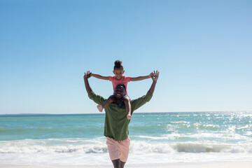 Cheerful african american father carrying daughter on shoulders at beach on sunny day, copy space - Powered by Adobe