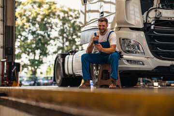 A bottom view of a mechanic drinking tea or coffee from a cup and sitting leaning against a truck in a workshop. A young caucasian men mechanic. Truck service, repair. Rest and break at work. - obrazy, fototapety, plakaty