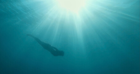 Beautiful underwater image freediver woman silhouette swimming from surface with sun rays to depth of blue sea. Unfocused image