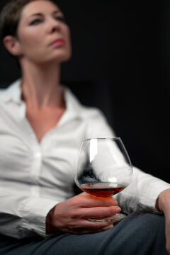 Drinking glass filled with alcohol held by sad looking woman in business clothes sitting with her legs crossed. Woman's image blurred. Concept for depression, sadness, alcoholism, drinking problems.