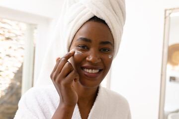 Portrait of smiling young african american woman applying moisturizer on cheeks in bathroom