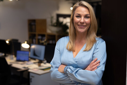 Portrait of smiling confident female professional with arms crossed standing in office at night