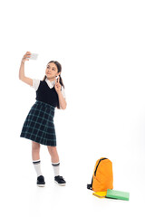 Schoolkid showing peace sign while taking selfie on smartphone near books and backpack on white background.