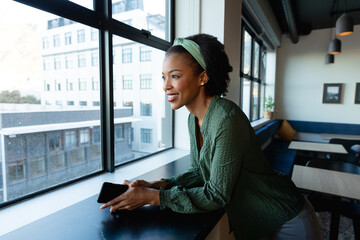 Smiling african american young businesswoman with smart phone looking through window in office
