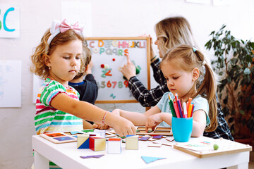 Little girls playing with cubes and geometric figures on desk in playroom, educator with boy...