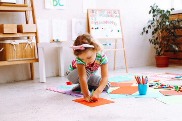 Little girl with nice bezel on head sitting on floor and drawing with colored pencils on sheet of...