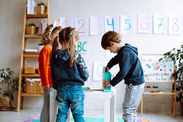 Little girls looking at boy opening box with toys on desk in playroom back view. Interesting game for kindergartners developing logic and intelligence. Communication with peers in kindergarten - obrazy, fototapety, plakaty