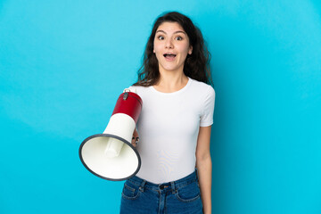 Teenager Russian girl isolated on blue background holding a megaphone and with surprise expression