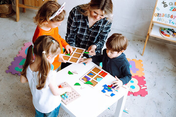 Little kids with educator folding colorful details of constructor on desk in playroom from above view. Interesting game for kindergartners developing logic and intelligence. Communication with peers - obrazy, fototapety, plakaty