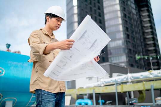 Asian Engineer With White Safety Helmet Hold And Look The Blue Print Paper Of Construction Site Also Stay On Terrace Or Deck Of The Building.