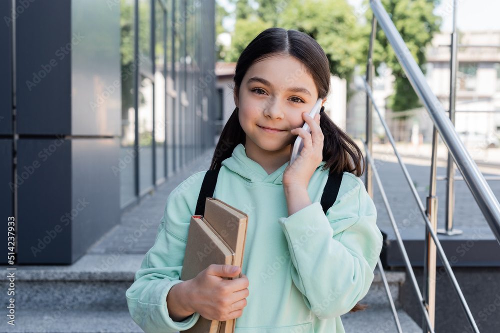 Sticker Portrait of pupil with books talking on cellphone on urban street.