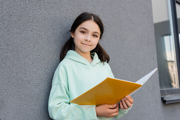 Smiling schoolkid holding notebook and looking at camera near building outdoors.