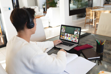 Asian teenage boy wearing headphones while learning online over laptop at home
