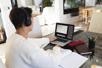 Obraz na płótnie Canvas Asian teenage boy with books and headphones studying online over video call on laptop at home