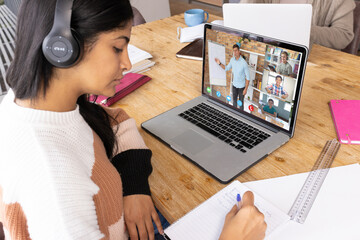 Serious biracial teenage girl writing in book while learning online over video call on laptop