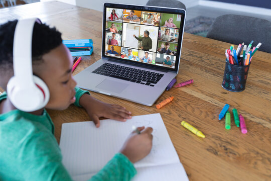 African american boy wearing headphones and listening to online class while writing in book at home