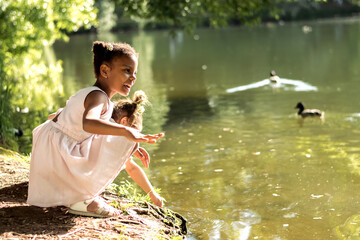 Happy caucasian and African-American girls watching birds on a pond on a summer day in the...