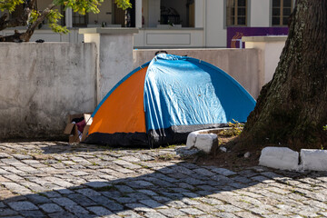 A tent on a city street near a tree. Homeless people in a tent live on the streets of the city. The...