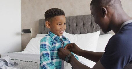 Video of happy african american father dressing up son in bedroom