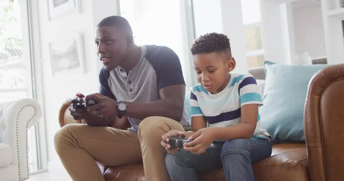 Video of happy african american father and son sitting on sofa and playing video games
